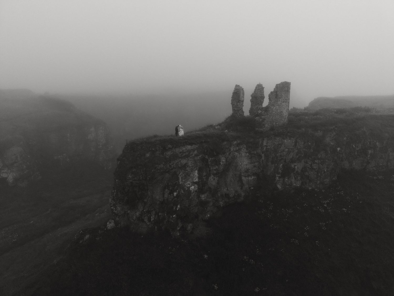 Black and white aerial view of ancient rock formations on a misty cliff edge with a lone figure standing nearby.