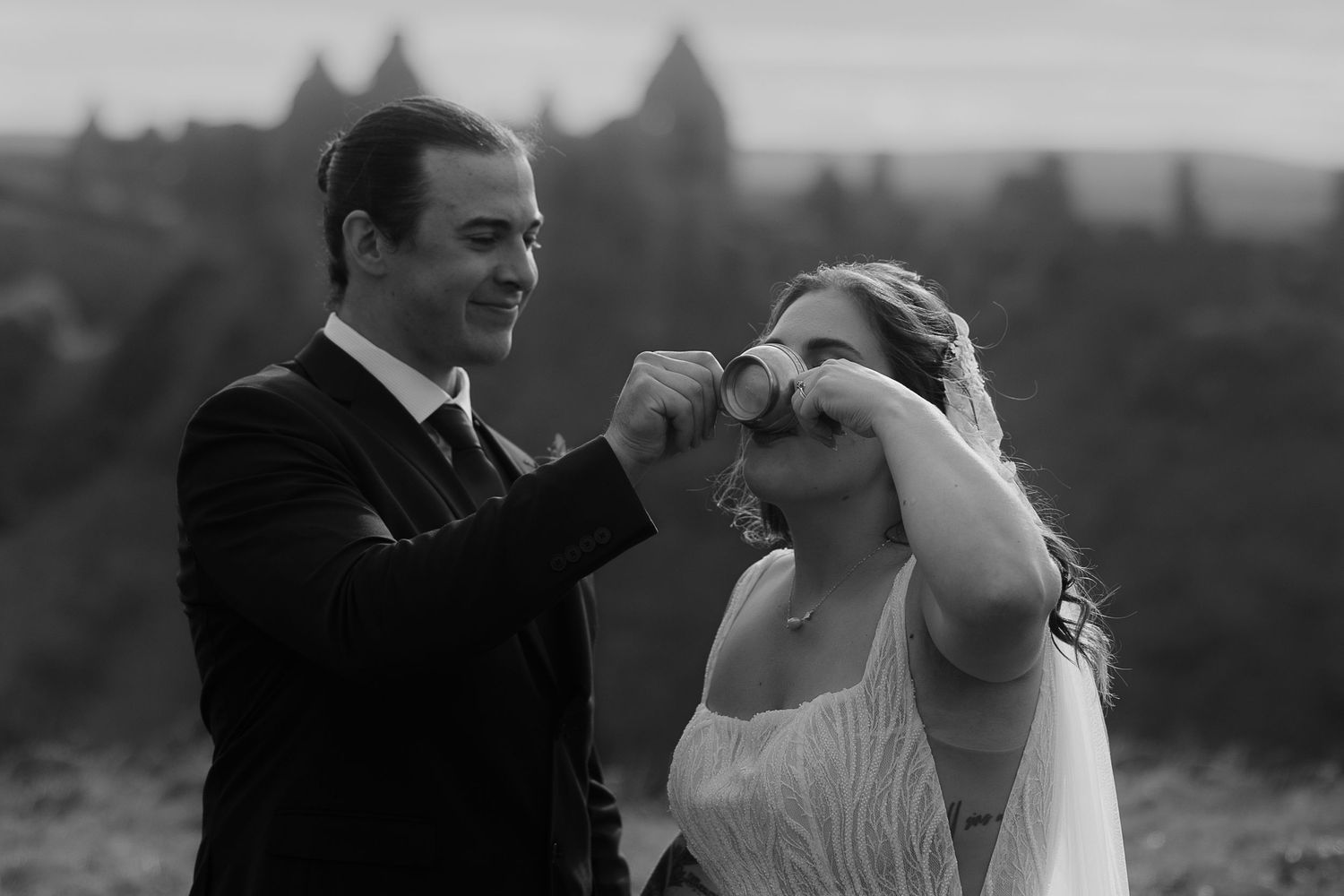 Black and white wedding photo of groom feeding bride a treat outdoors with historic architecture in background.