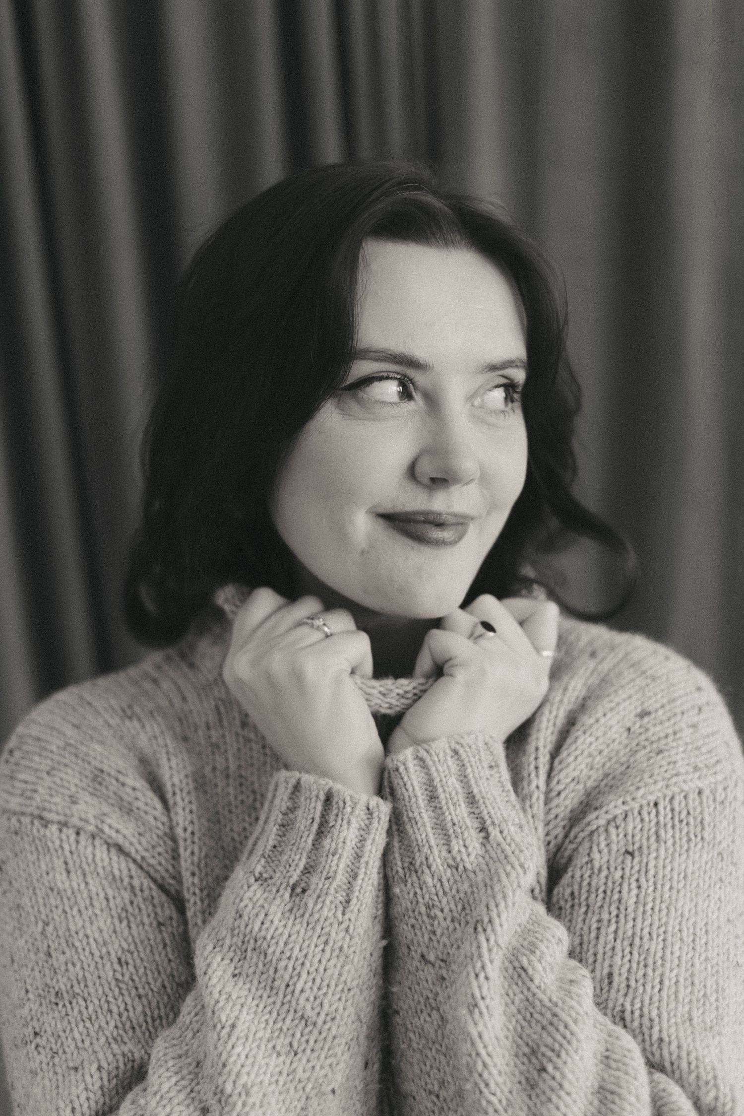 Black and white portrait of a smiling woman in a cozy knit sweater, hands near collar, looking up thoughtfully.