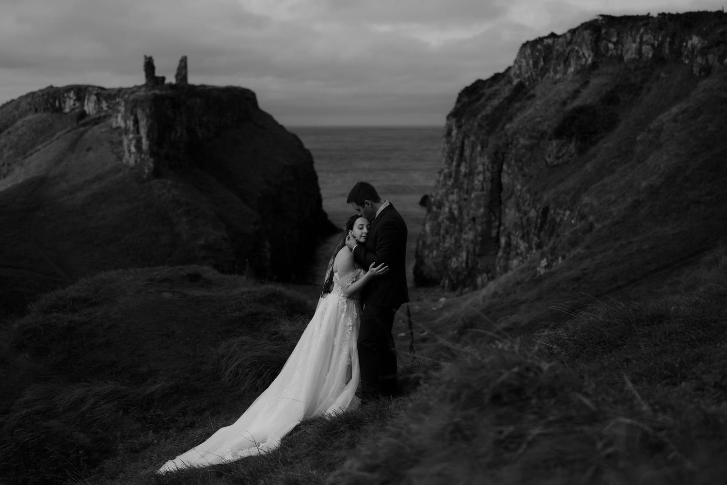 A wedding portrait on a dramatic coastal cliff with castle ruins in the background.