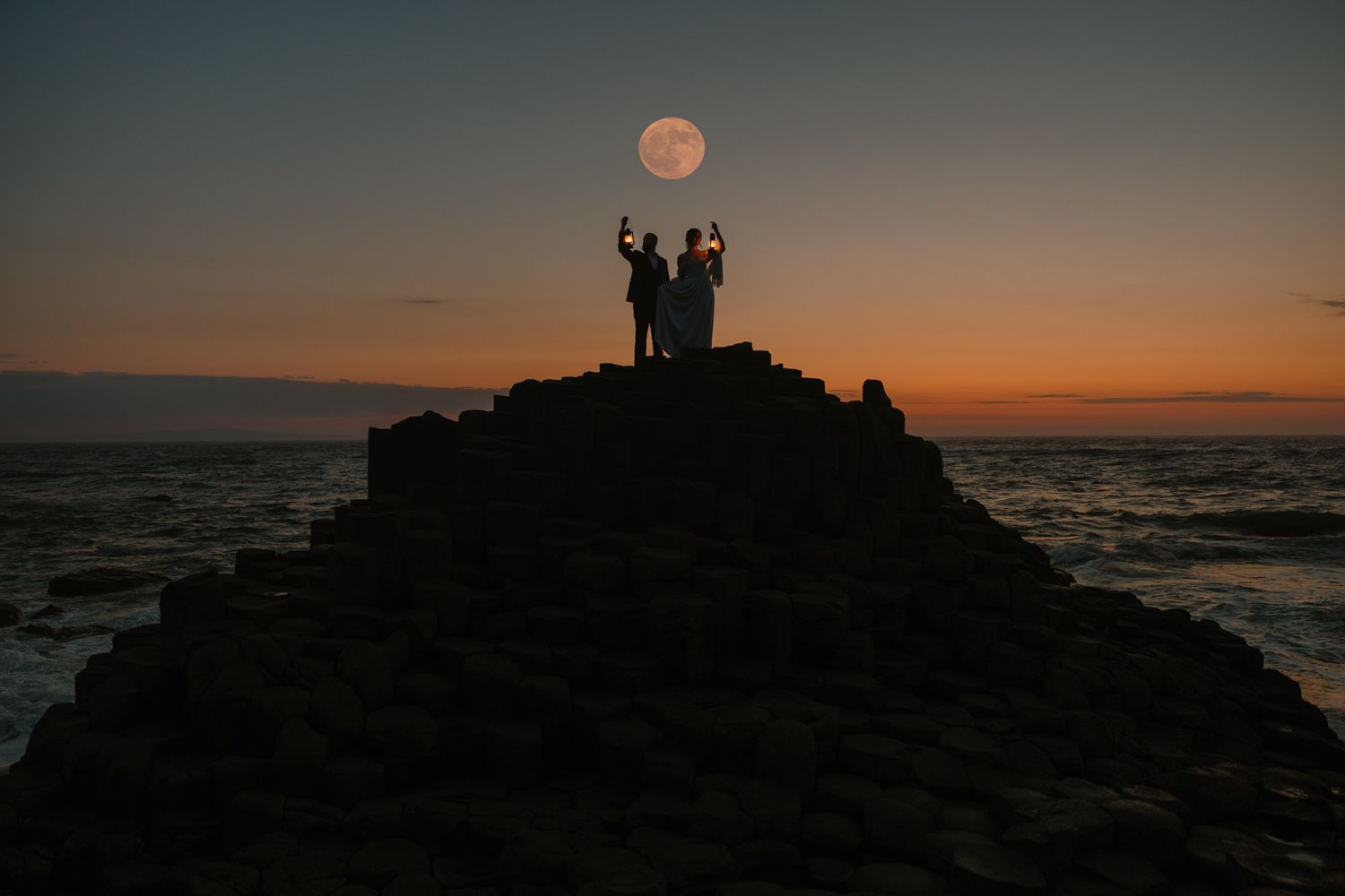 Silhouettes stand on rocky outcrop against vibrant orange sunset over ocean horizon.