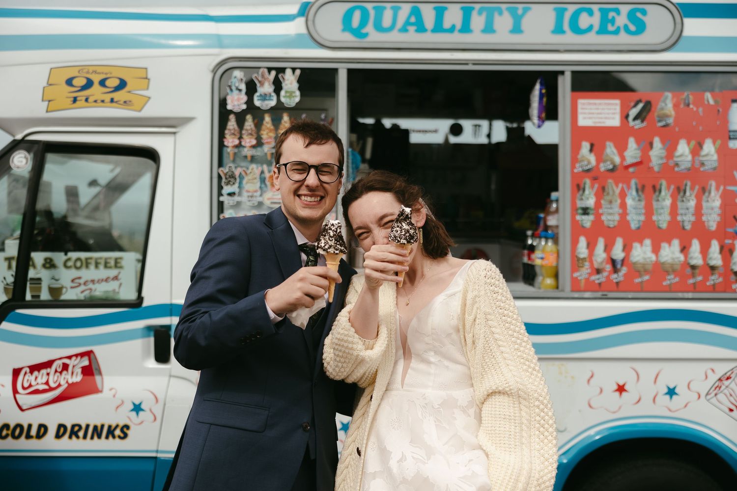 An ice cream truck with 'Quality Ices' branding serves customers on a sunny day while people gather around to order.