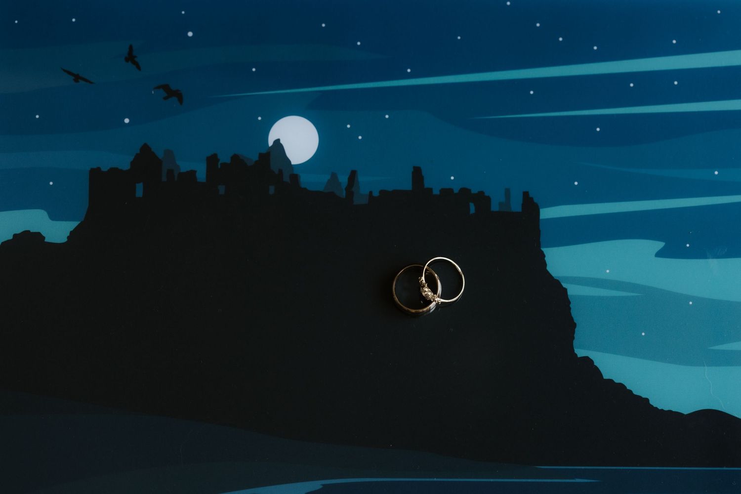 Moonlit castle silhouette on a rocky cliff at night with two gold wedding rings and stars in the sky.