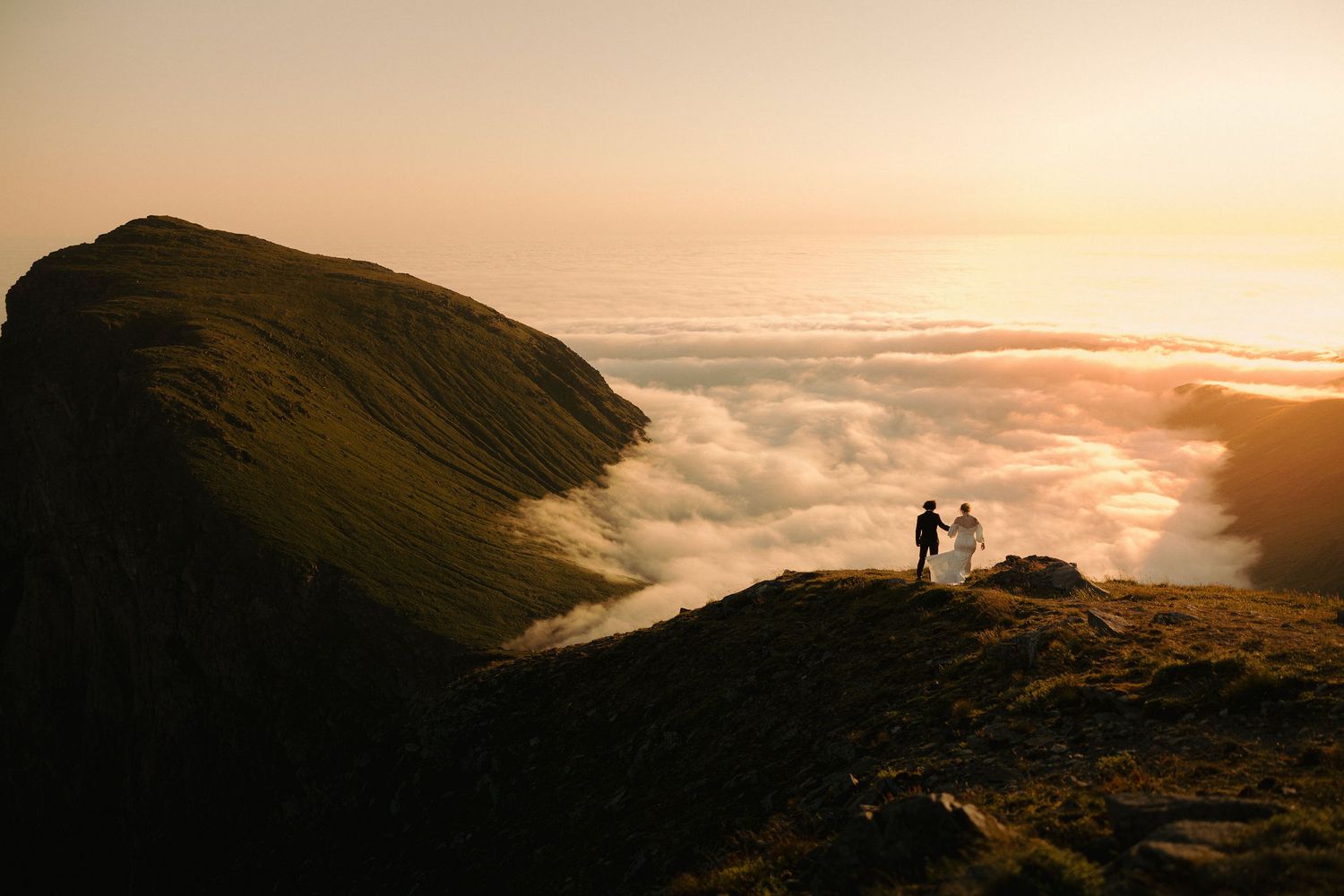 Silhouetted couple stands on mountain peak overlooking dramatic sunset and sea of clouds in misty landscape.