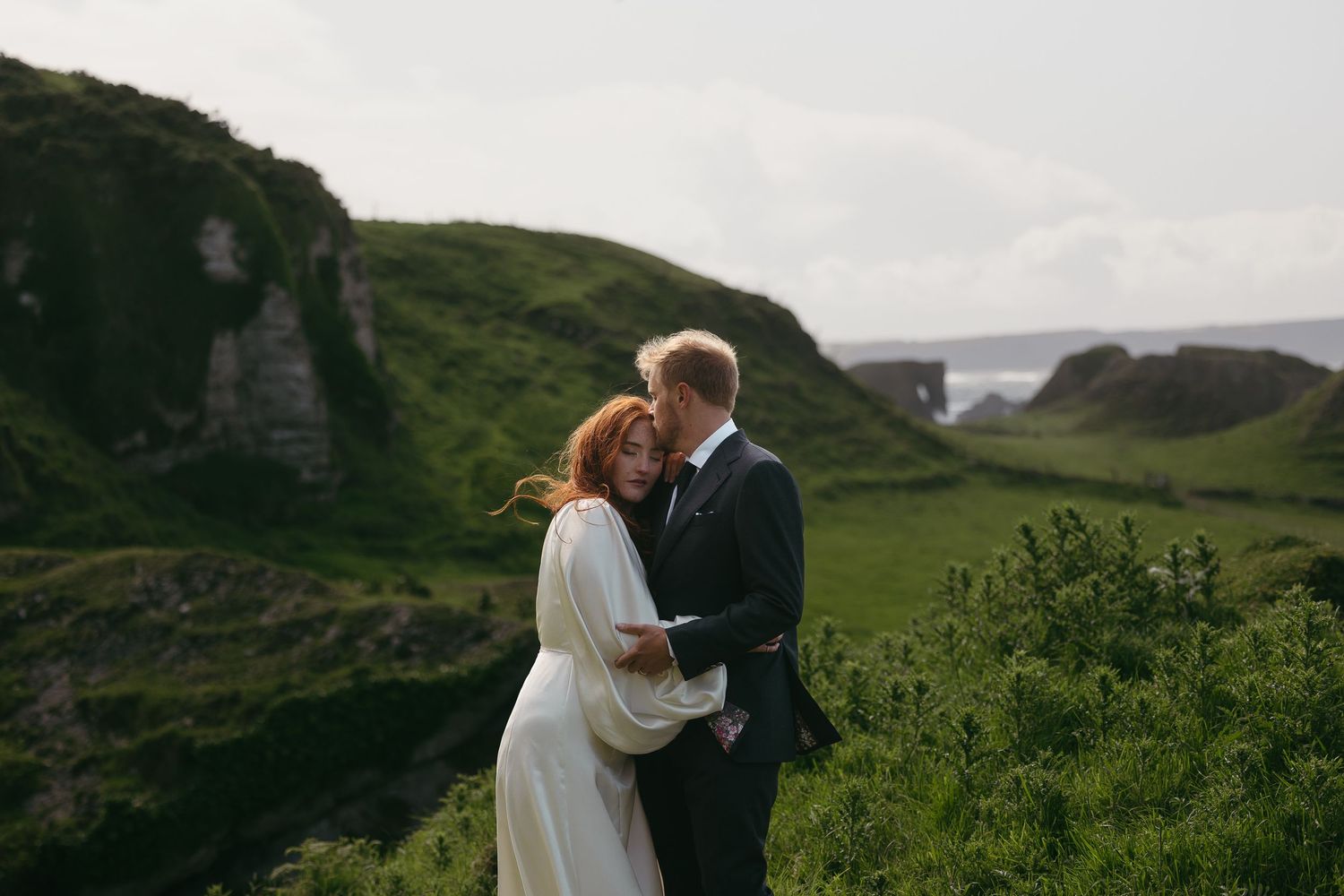 A romantic wedding photo against dramatic coastal cliffs with a natural stone arch formation in the background.