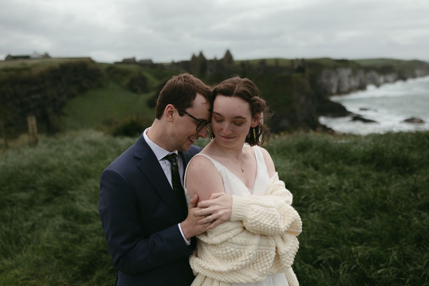 A couple embraces on a green coastal hillside overlooking the ocean on an overcast day.