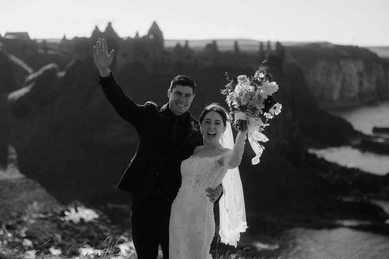 Black and white photo of a joyful bride and groom celebrating on a dramatic cliff edge with stunning rock formations.