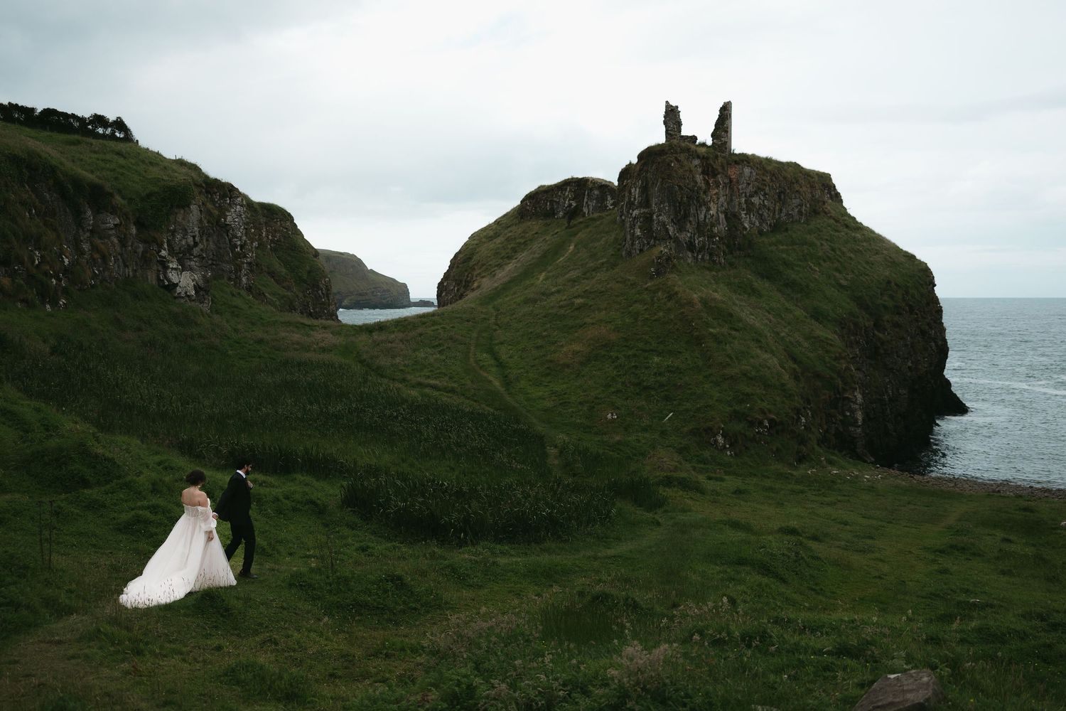 Couple walking near ancient castle ruins on dramatic coastal cliffs.
