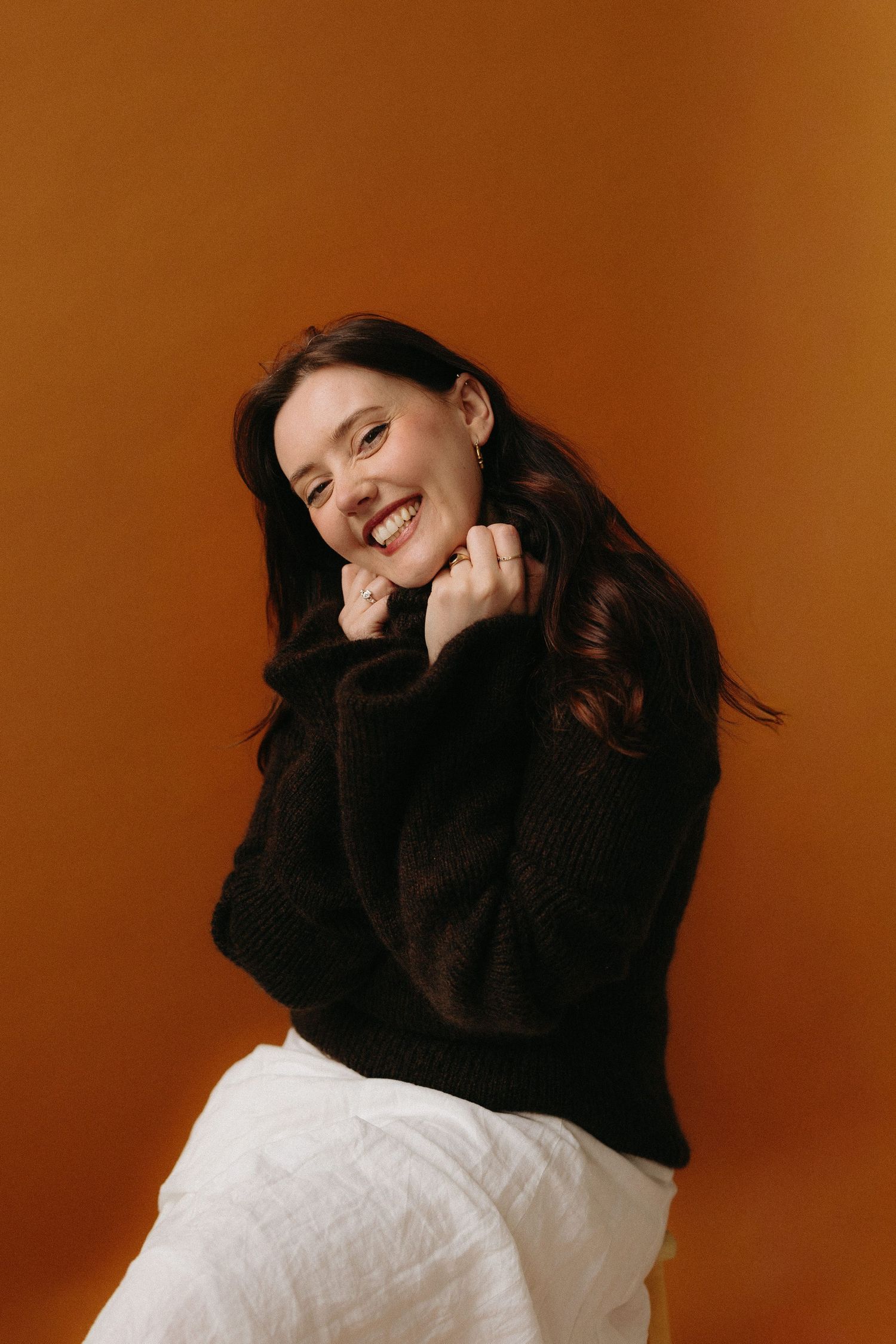 Smiling brunette woman in black knit sweater and white skirt posing against warm orange background.