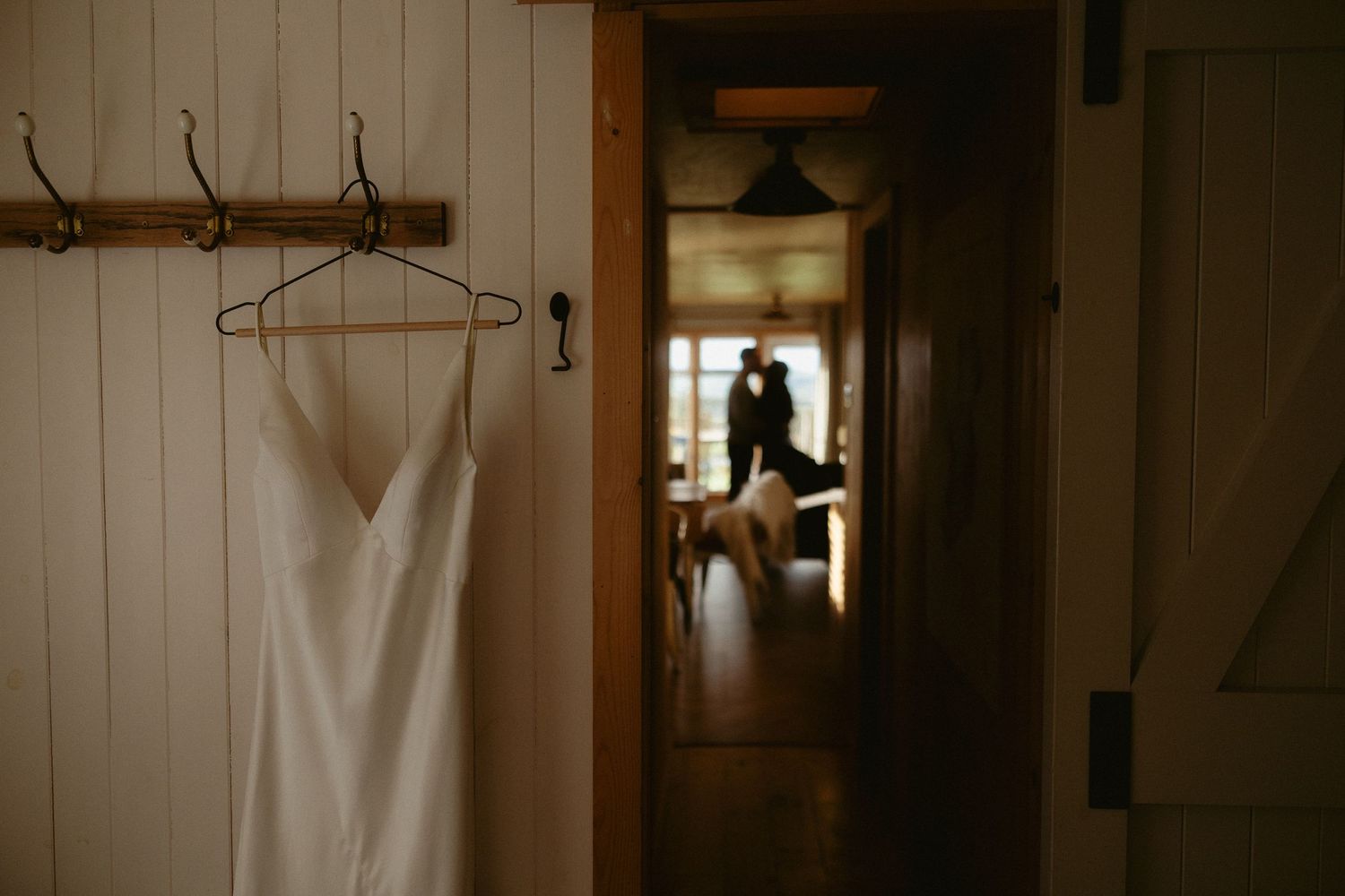 White wedding dress hanging on wall hook with couple silhouetted in background hallway of rustic venue.