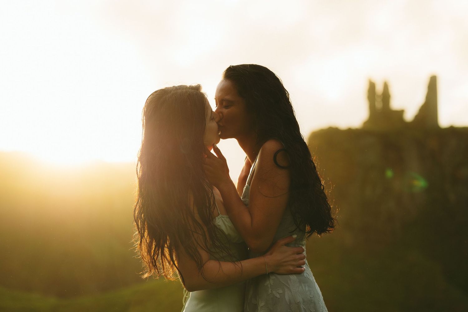 Two women sharing a romantic kiss at sunset with warm golden light and ruins in the background.
