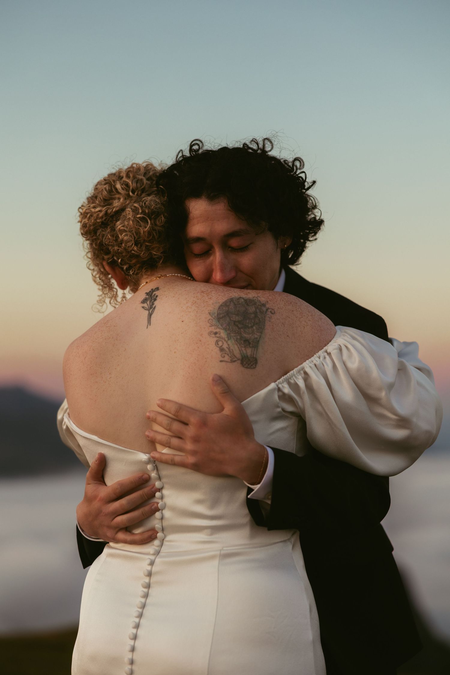 Intimate embrace captured against soft sunset lighting on coastal backdrop.