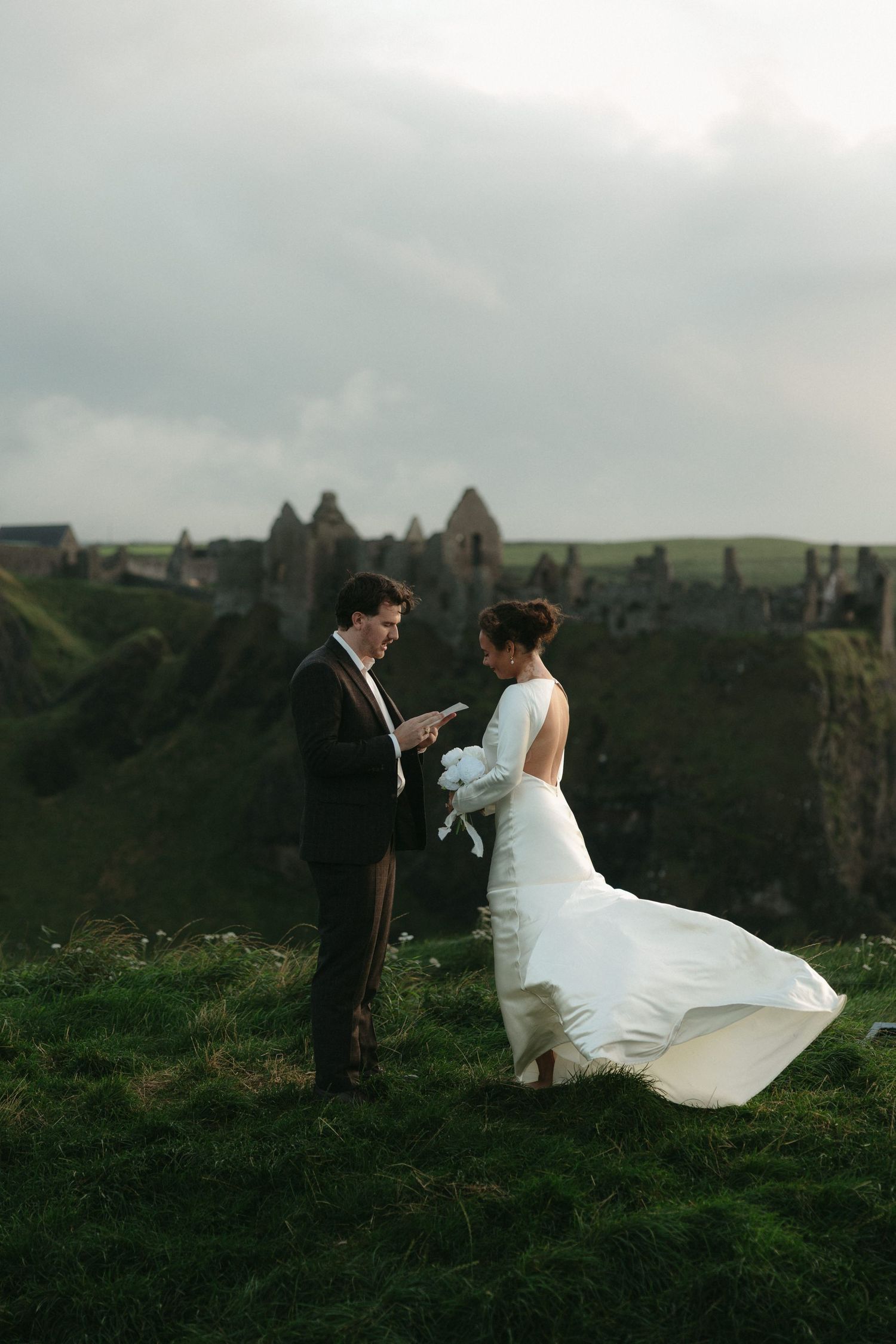A couple in wedding attire stands on a misty hilltop overlooking dramatic green landscapes and ancient ruins.