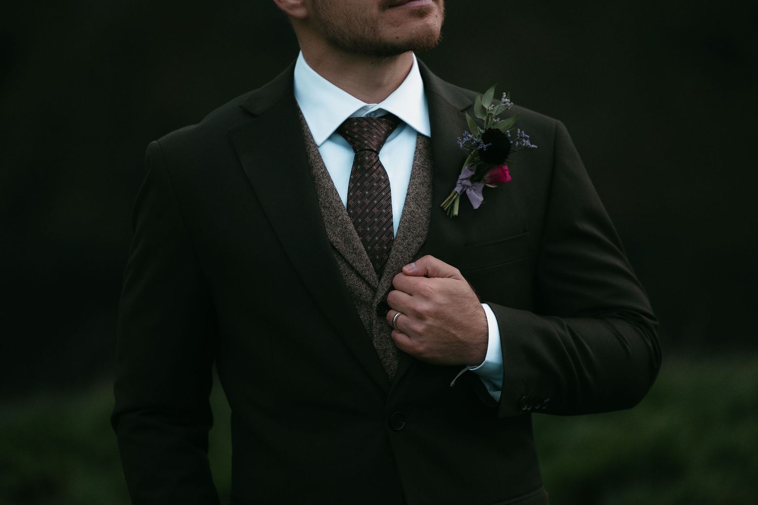 Groom in dark suit with floral boutonniere adjusting his tie outdoors.