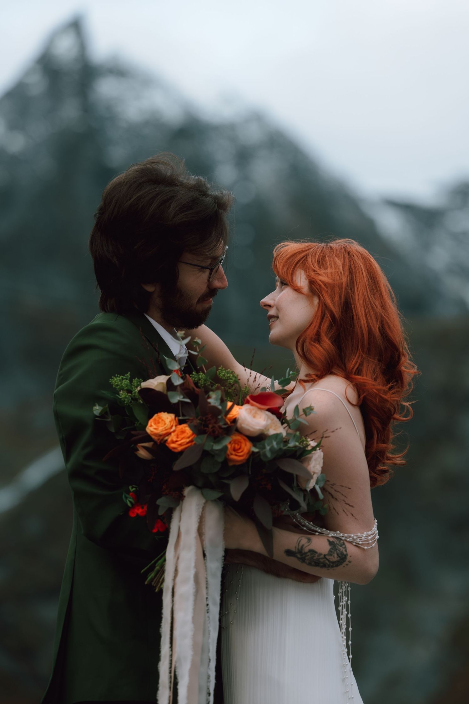 Wedding couple sharing intimate moment with floral bouquet against mountain backdrop.