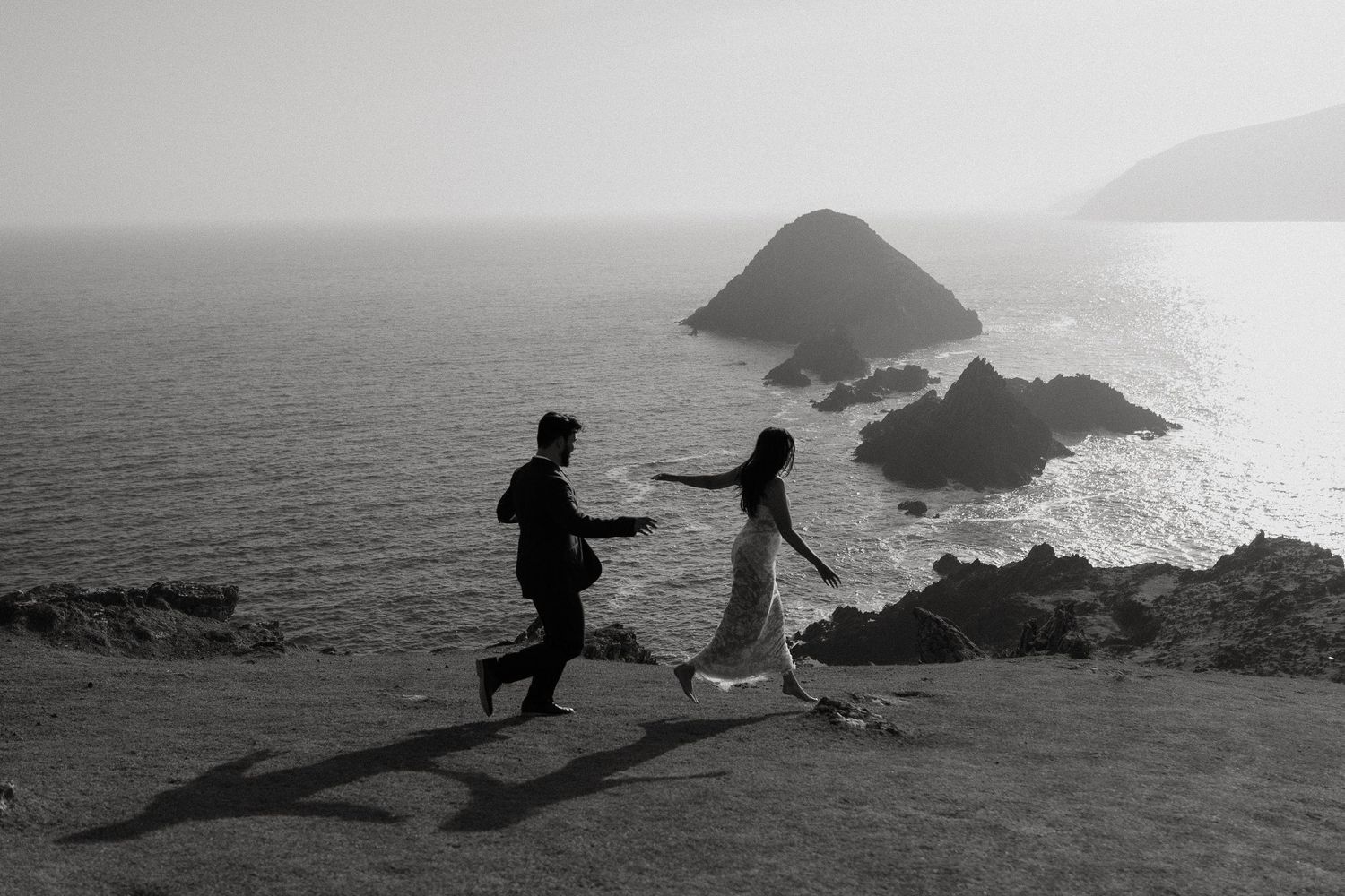 Black and white photo of a couple running on a cliff overlooking the ocean with rocky islands in the background.