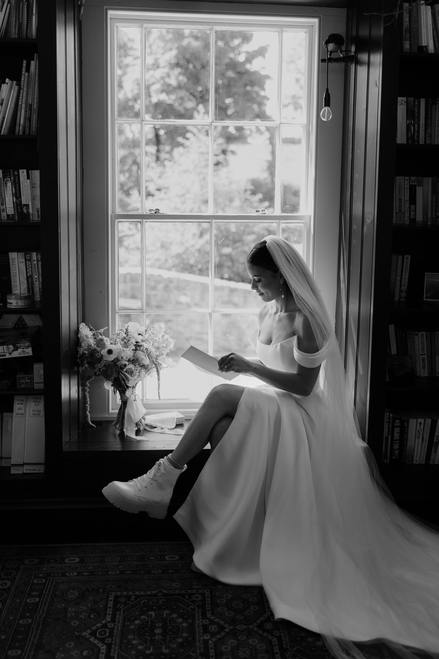 Black and white photo of a bride in a wedding dress and veil sitting by a window reading a letter in a library.