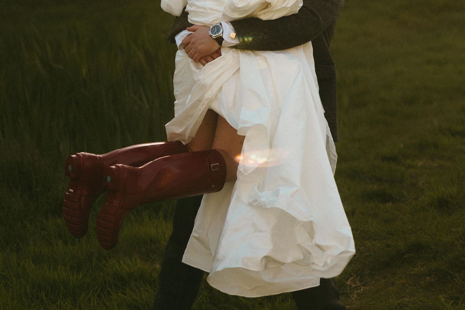 Groom carrying bride in white dress and red cowboy boots on green grass at rustic outdoor wedding.