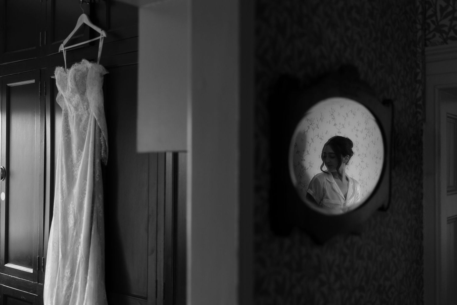 Black and white photo of a wedding dress hanging while a bride prepares, reflected in a vintage round mirror.