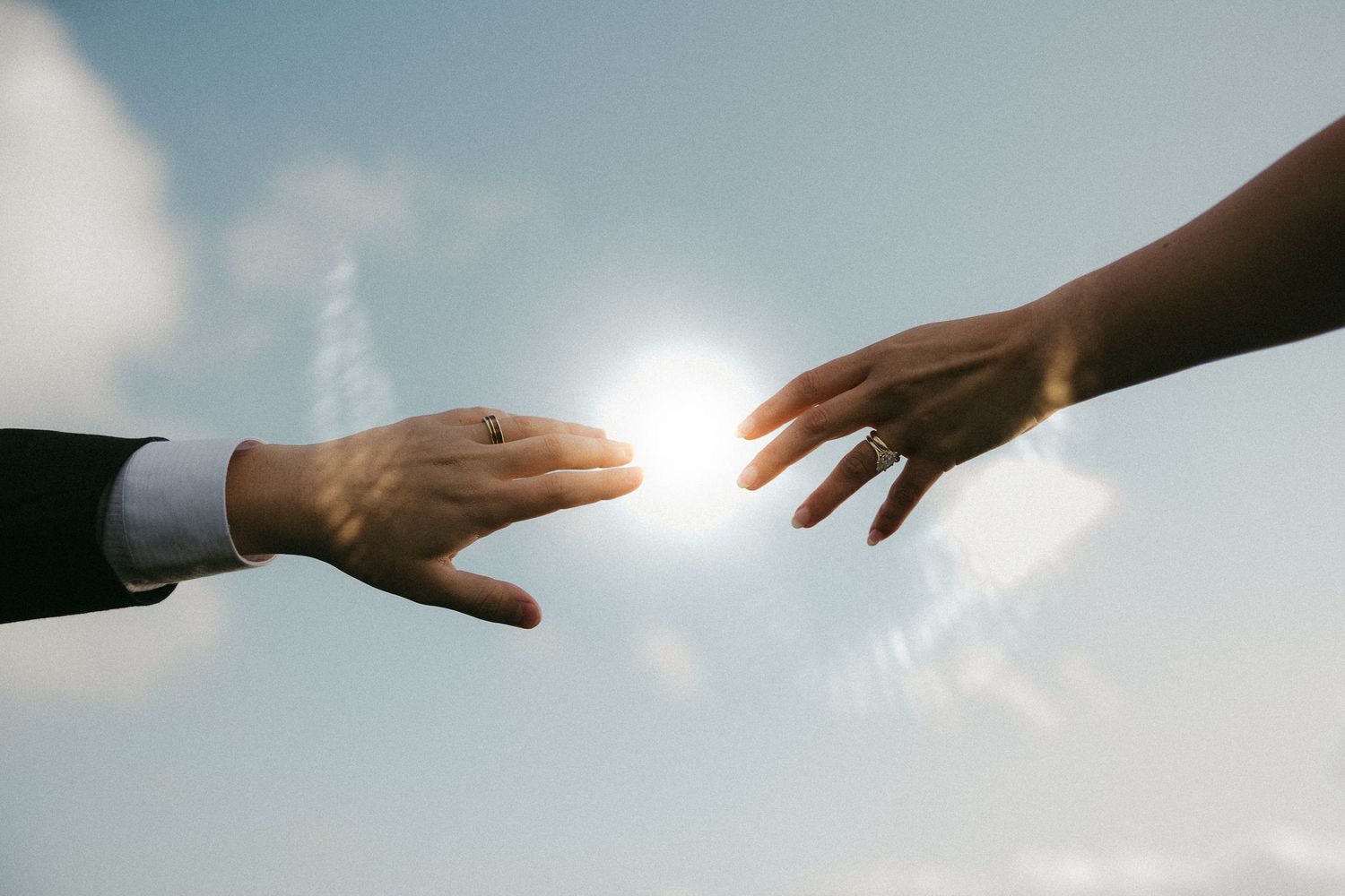 Hands reaching toward bright sunlight against a blue sky with wispy clouds.