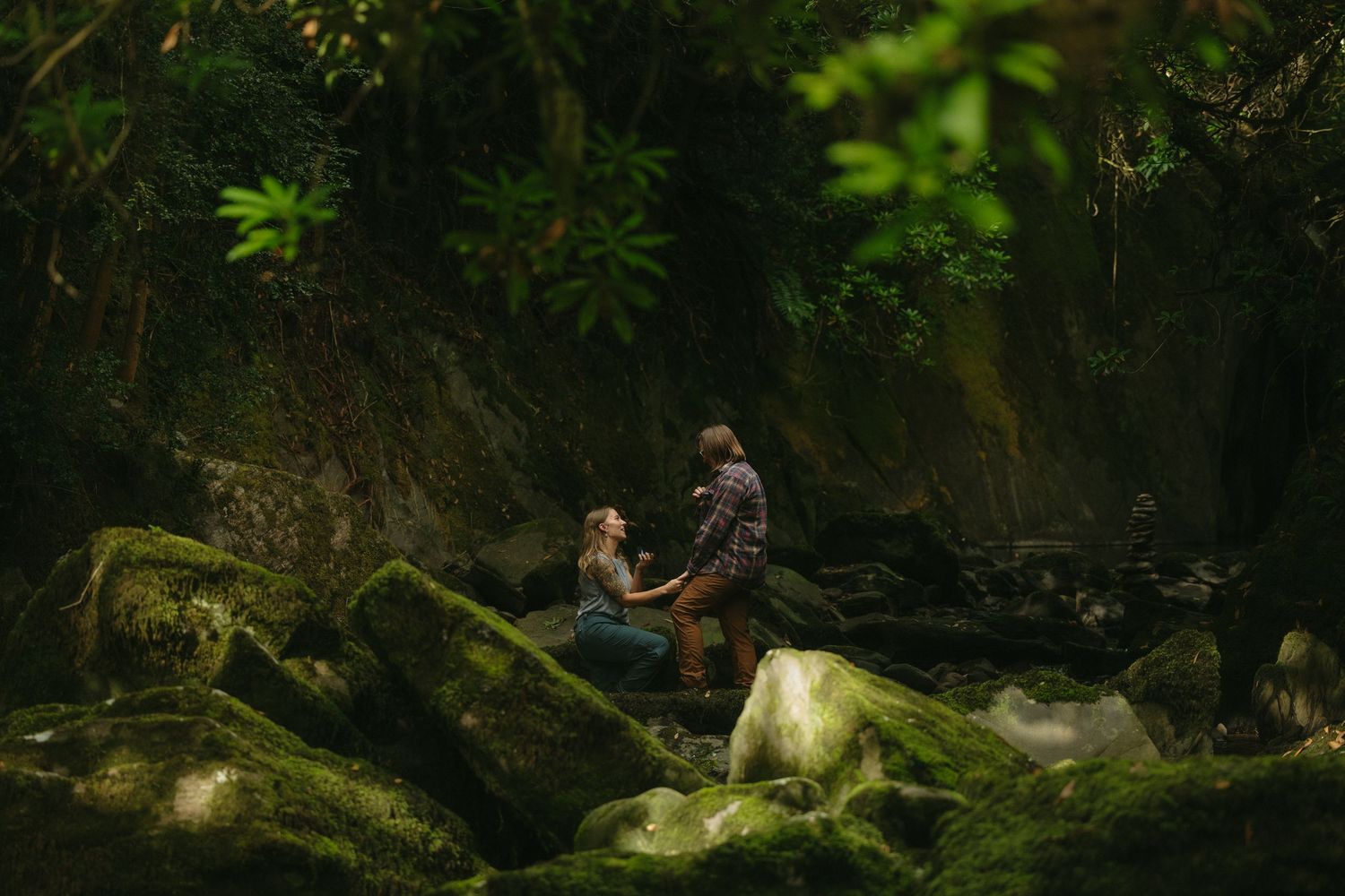 Couple sharing a romantic moment on moss-covered rocks beside a stream in a lush, green forest setting.