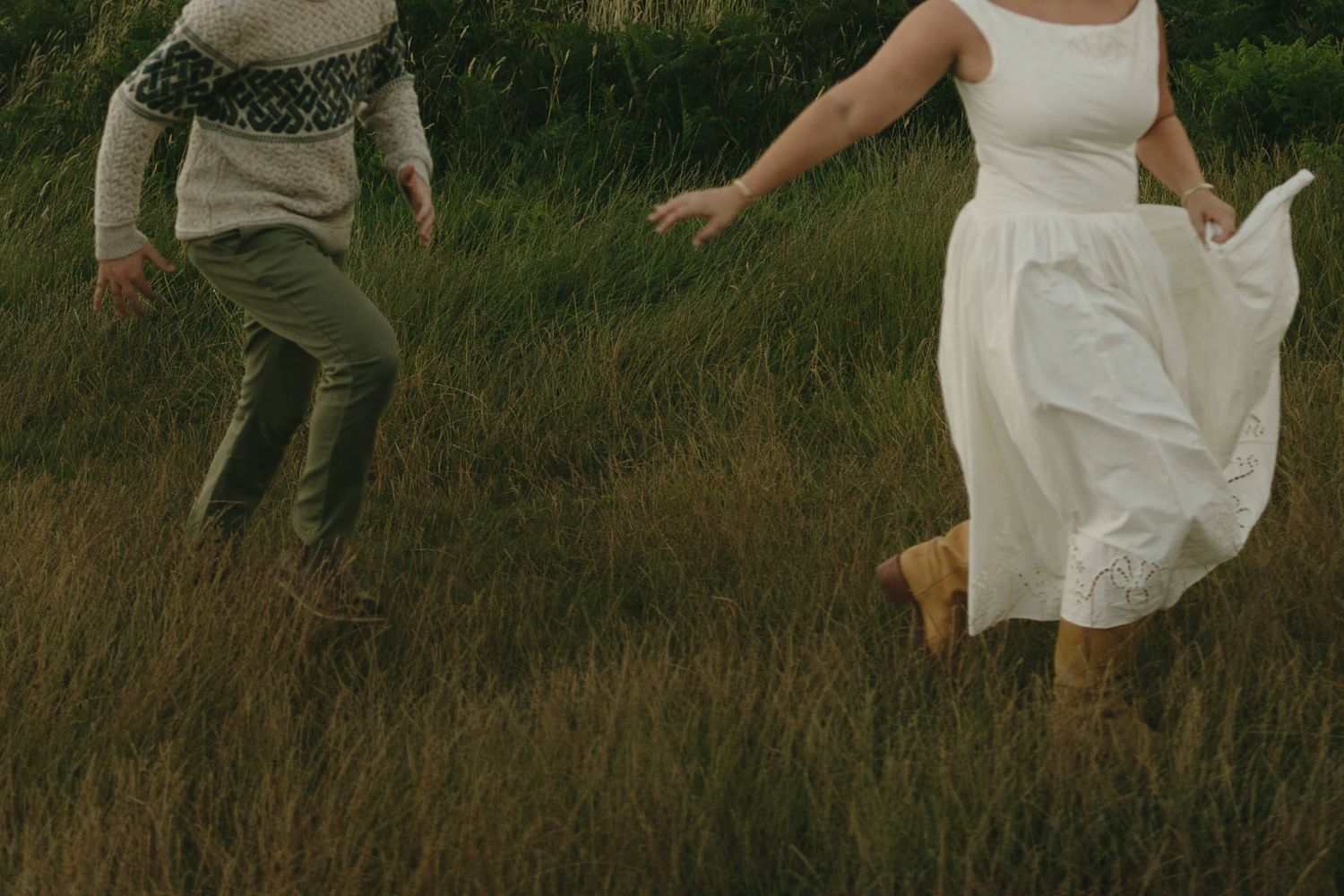 Couple running joyfully through tall green grass, woman in white dress and man in sweater and green pants.