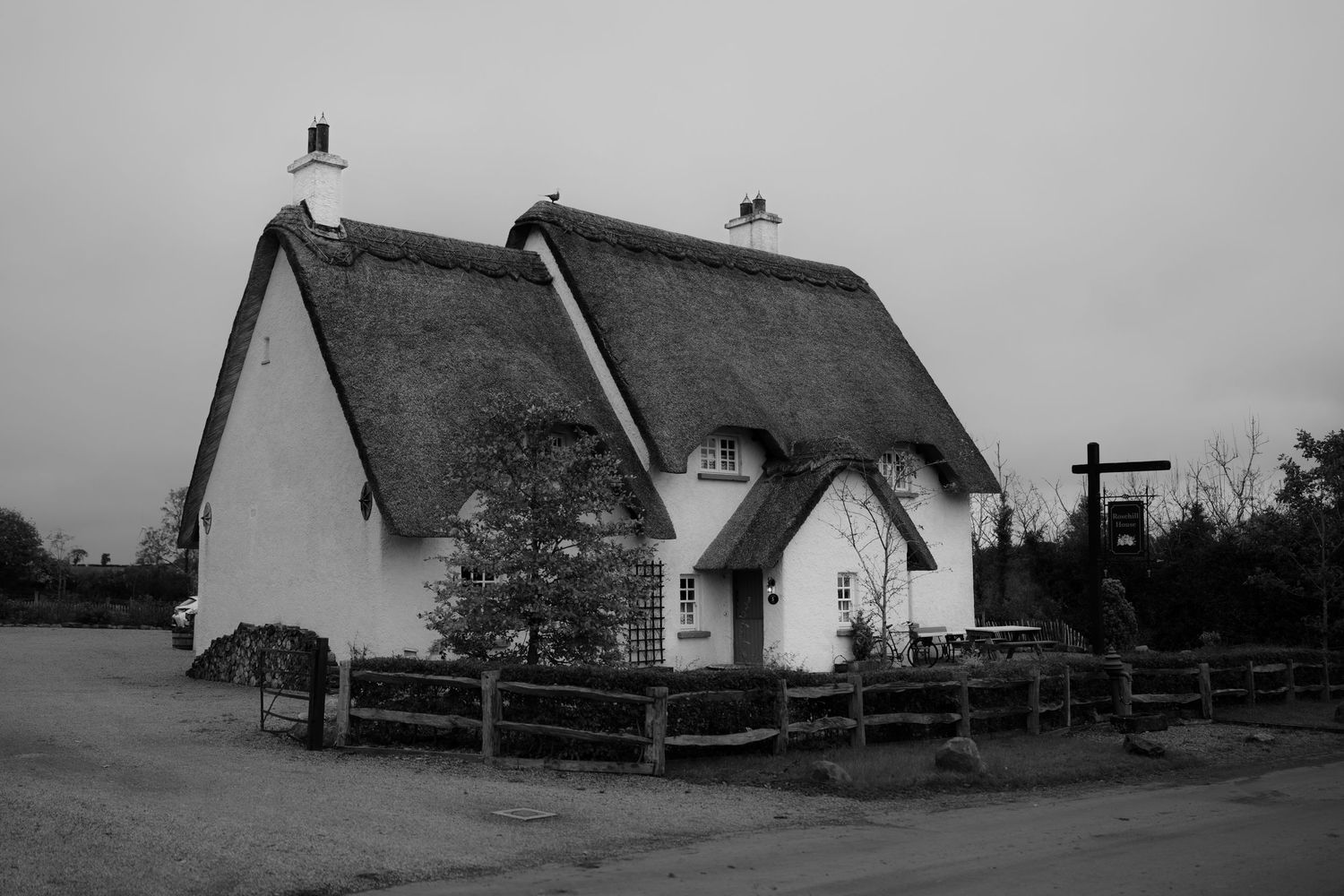 Black and white photo of traditional English thatched cottages with white walls and wooden fence on overcast day.
