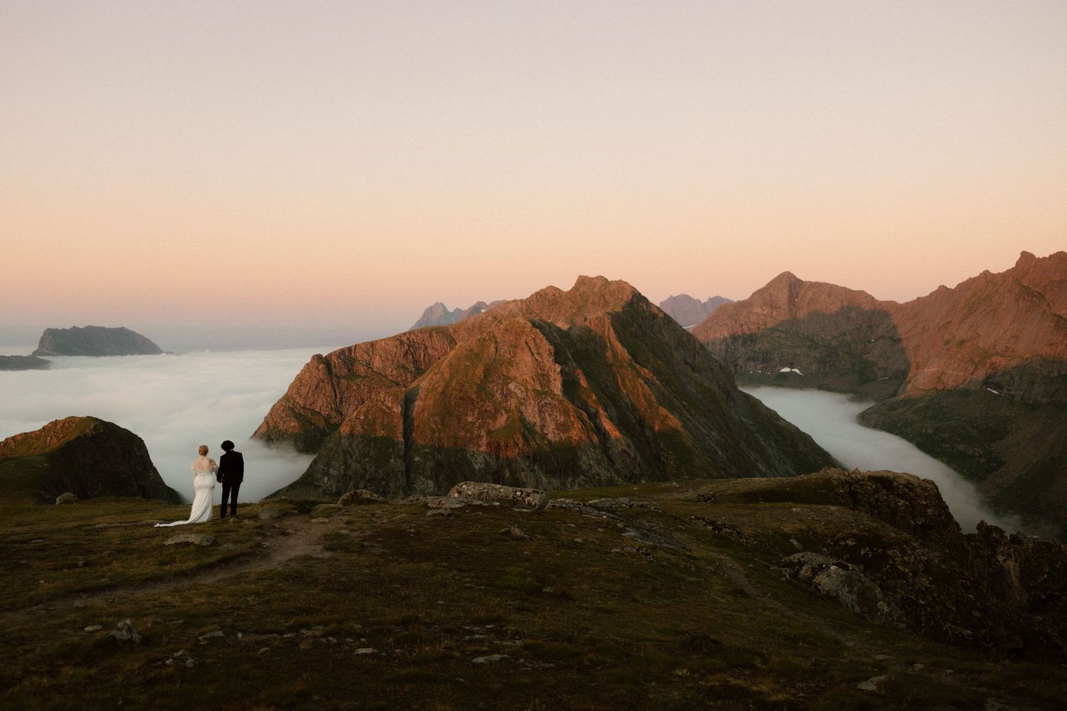 Silhouetted figures stand on rocky coastal cliffs at sunset overlooking dramatic mountain peaks and ocean views.