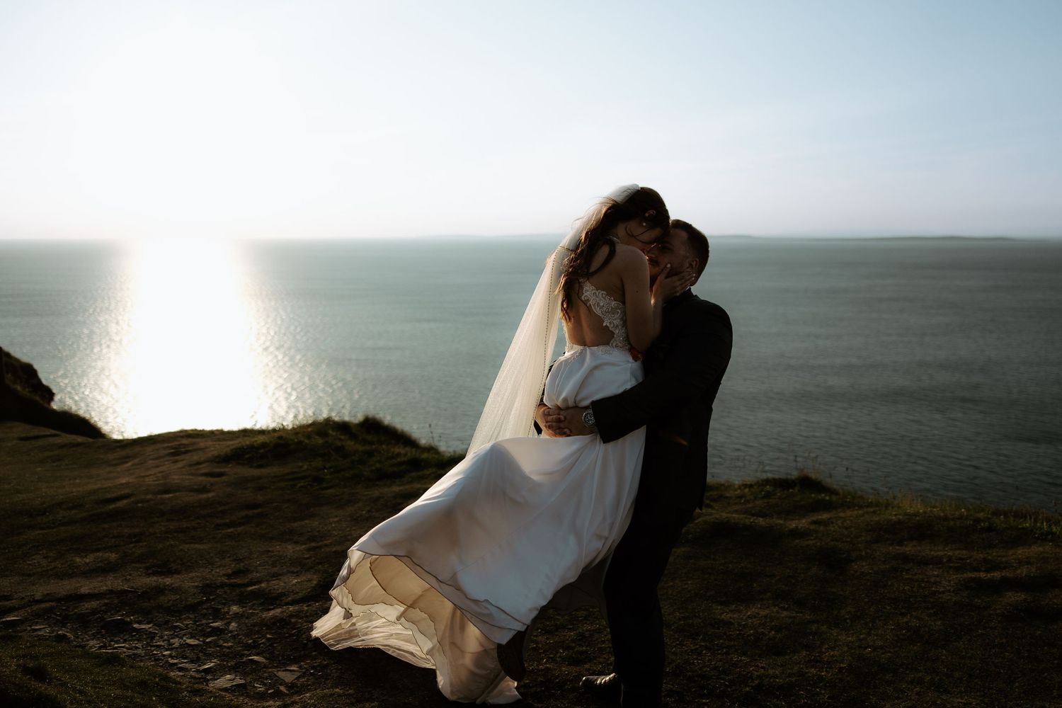 A silhouetted couple embraces on a cliff edge at sunset over the ocean.