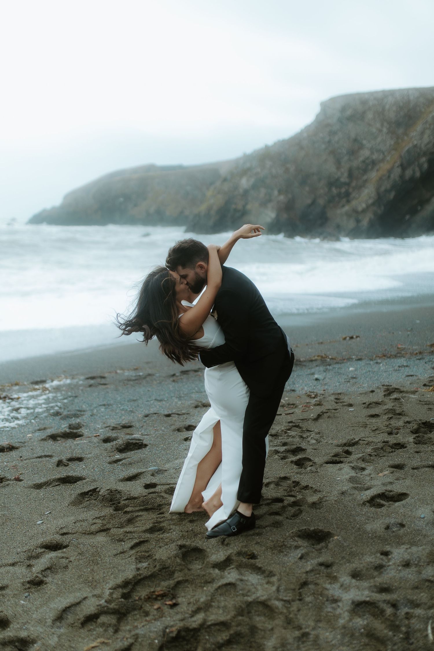 Couple sharing a romantic dip kiss on a dark sandy beach with dramatic ocean waves and misty cliffs behind them.