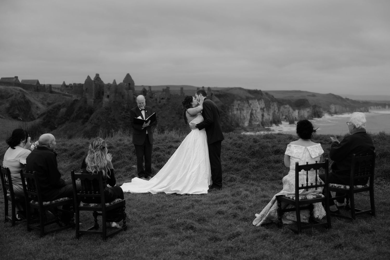 Black and white photo of a couple exchanging vows at a cliffside wedding ceremony with castle ruins and ocean in background.