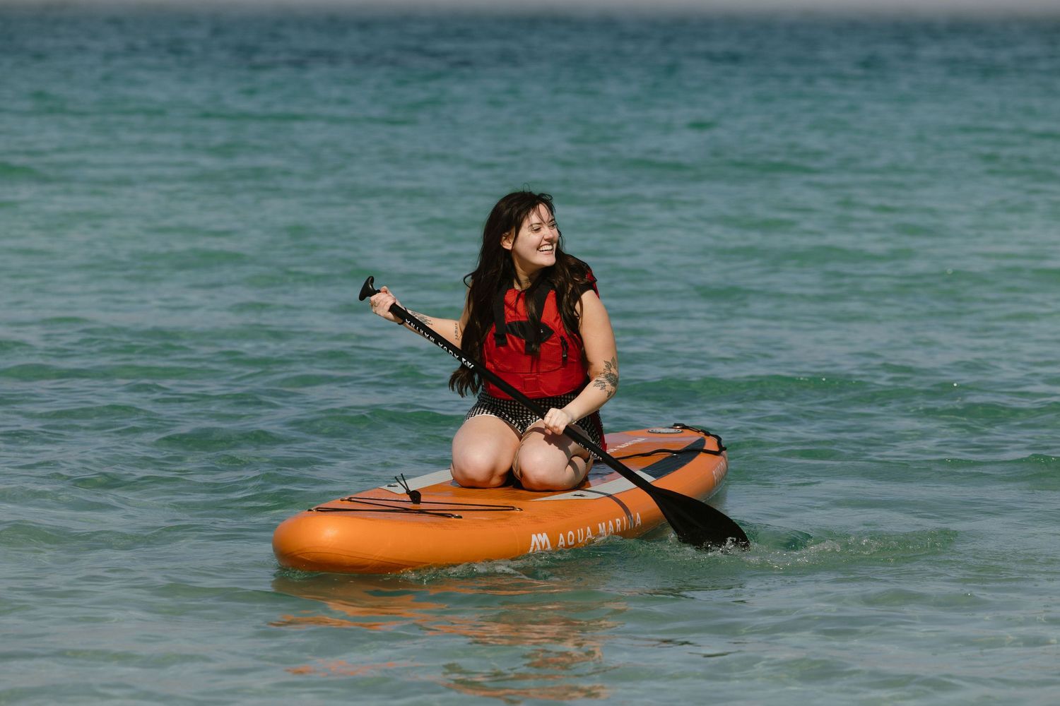 Person kayaking in bright orange inflatable boat on turquoise tropical waters.