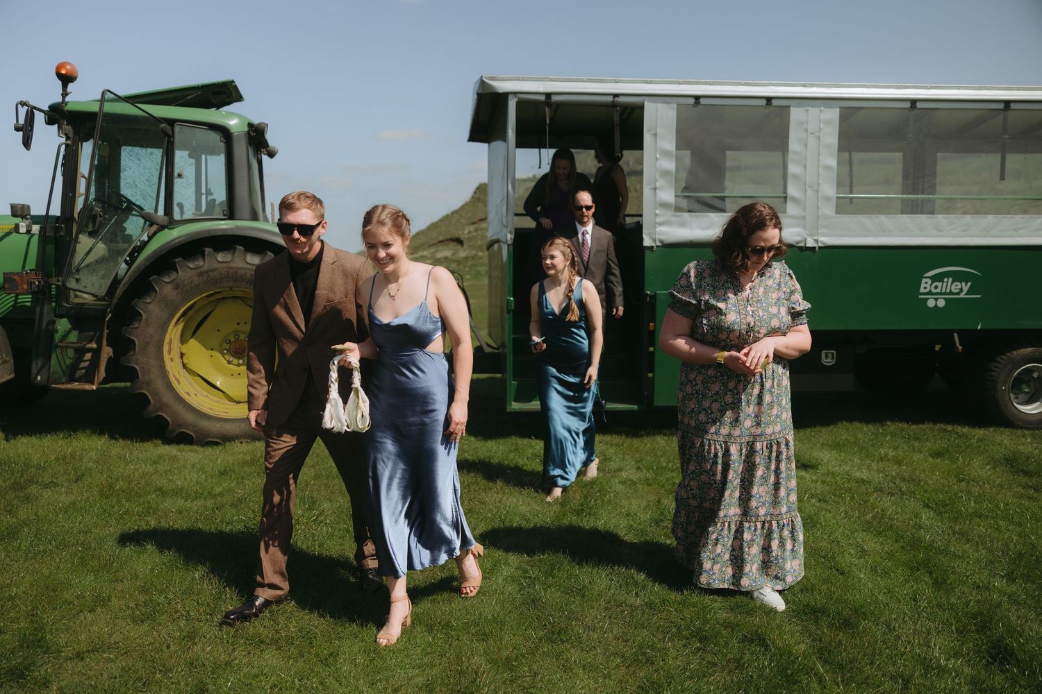 A group of people gather on a sunny lawn near green farm equipment and tractors during an outdoor event.