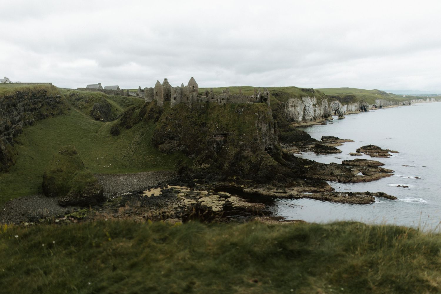 Ruins of Dunluce Castle on dramatic rocky cliffs along the Northern Ireland coastline under overcast skies.