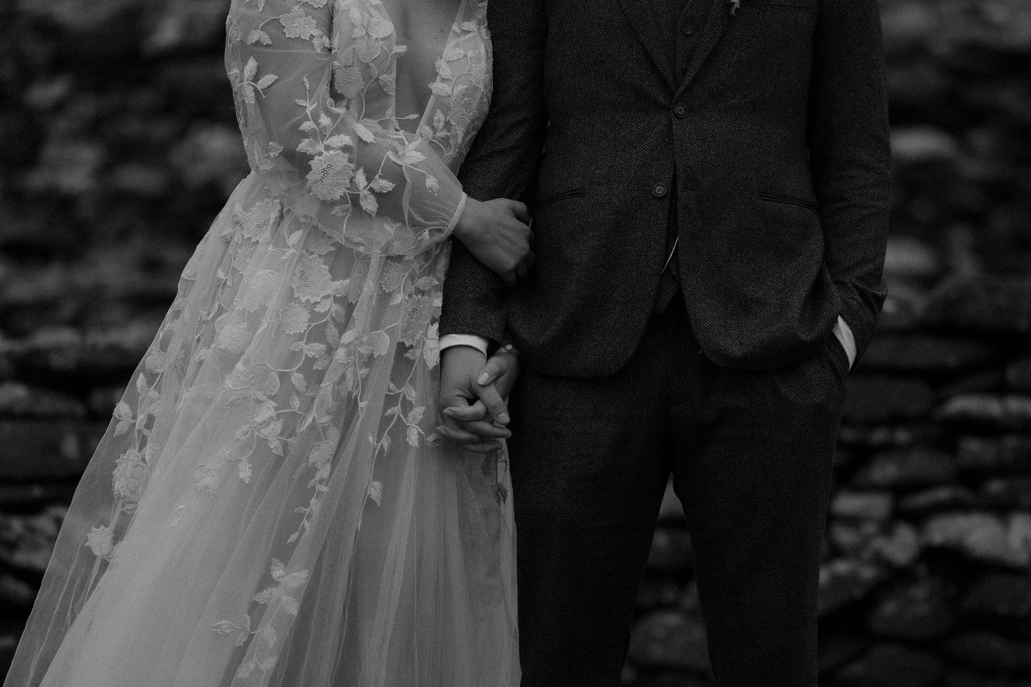 Black and white photo of a bride in floral lace gown holding hands with groom in dark suit at outdoor wedding.