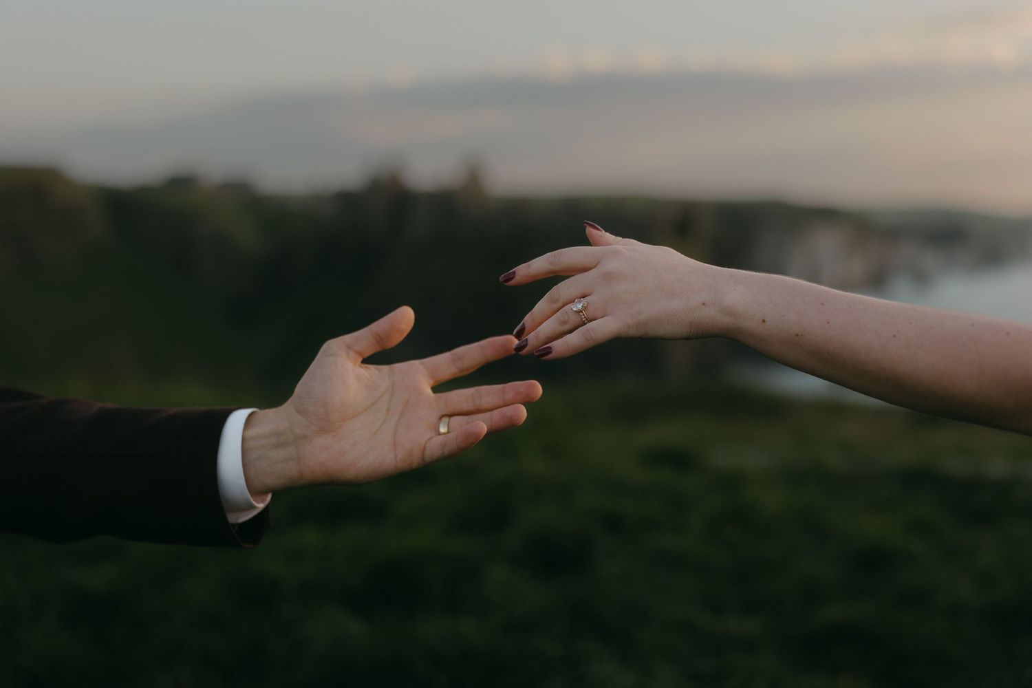 Two hands reaching toward each other at sunset, both wearing wedding rings, with a lush green landscape behind.