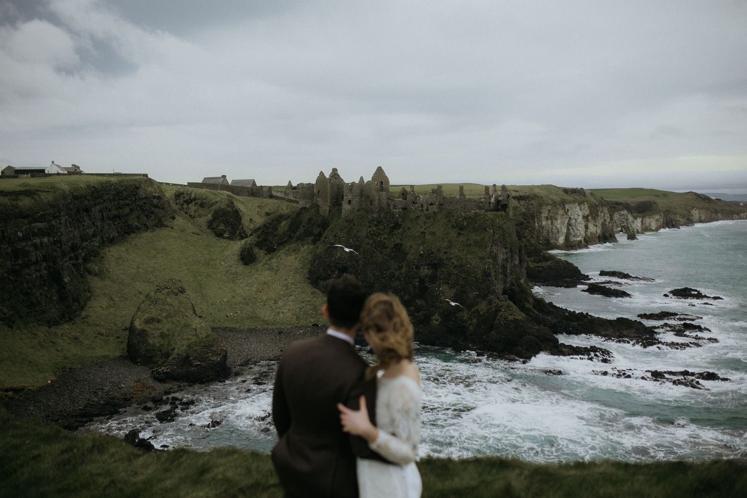 A couple embraces while overlooking dramatic coastal cliffs and crashing waves along a rugged shoreline in Ireland.