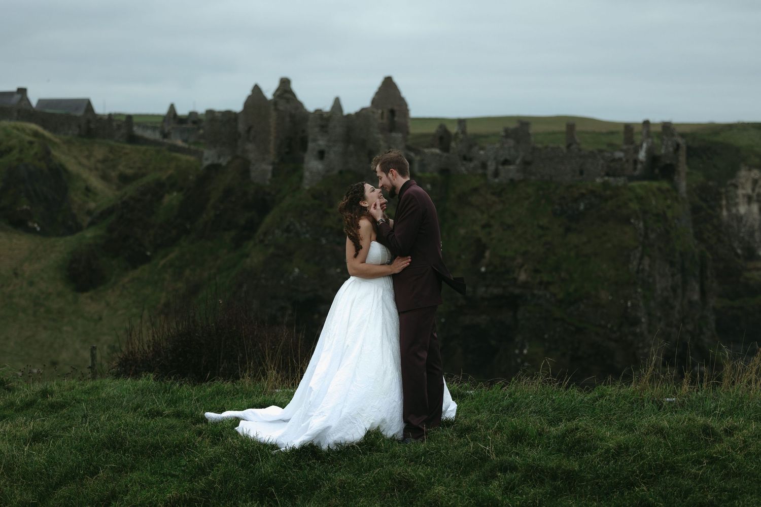 Bride and groom sharing romantic moment at Dunluce Castle ruins, Ireland, lush green hills backdrop.