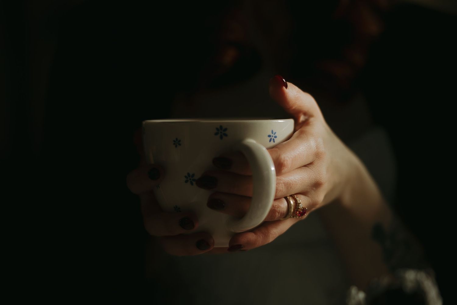 A woman with red nails and rings holds a white Christmas mug with snowflake patterns in a dark, moody setting.
