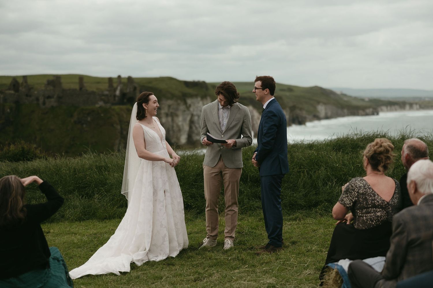 A wedding ceremony takes place on a scenic coastal cliff overlooking the ocean on a cloudy day.