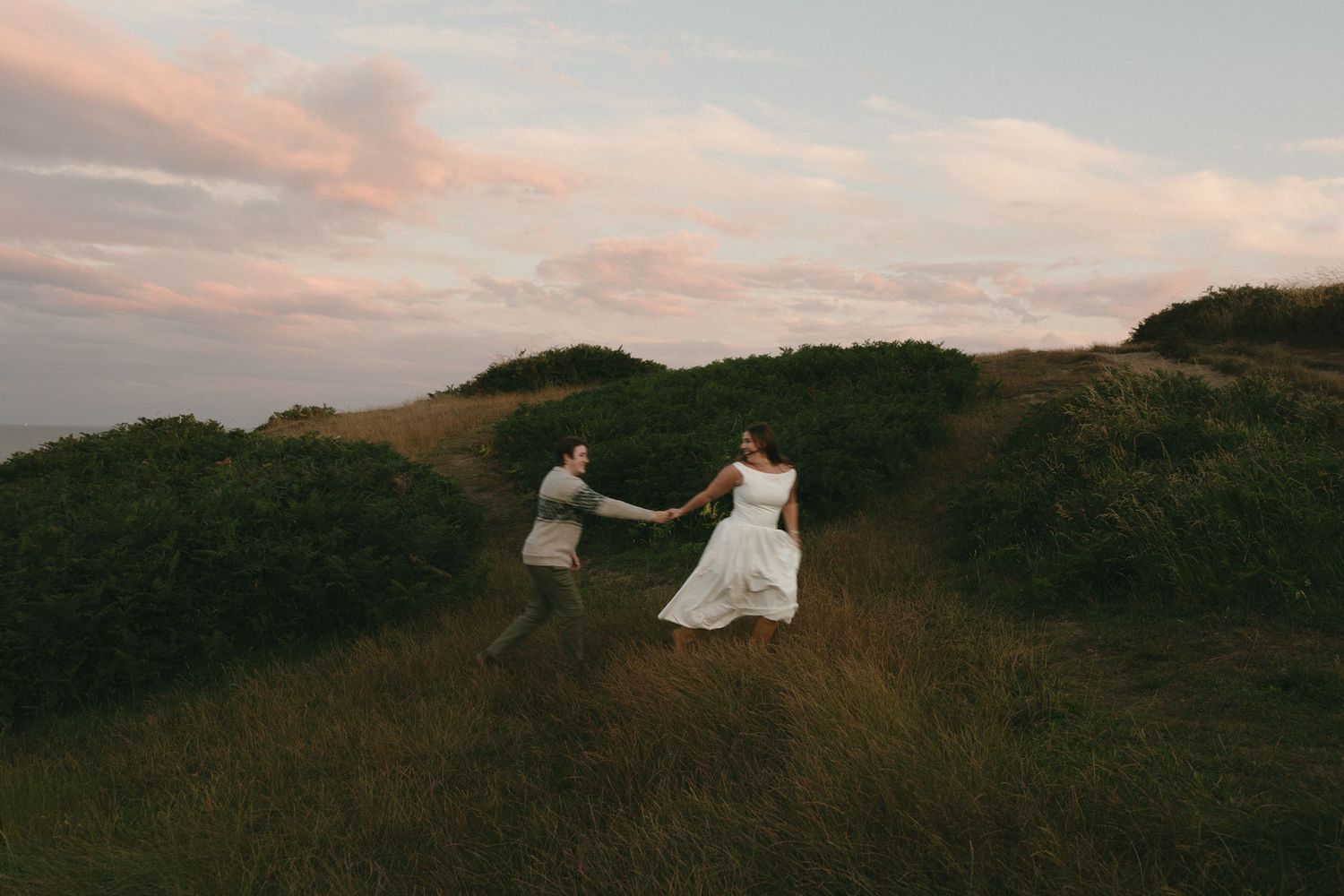 A bride and groom hold hands while walking through lush green hills at sunset during their romantic outdoor wedding.