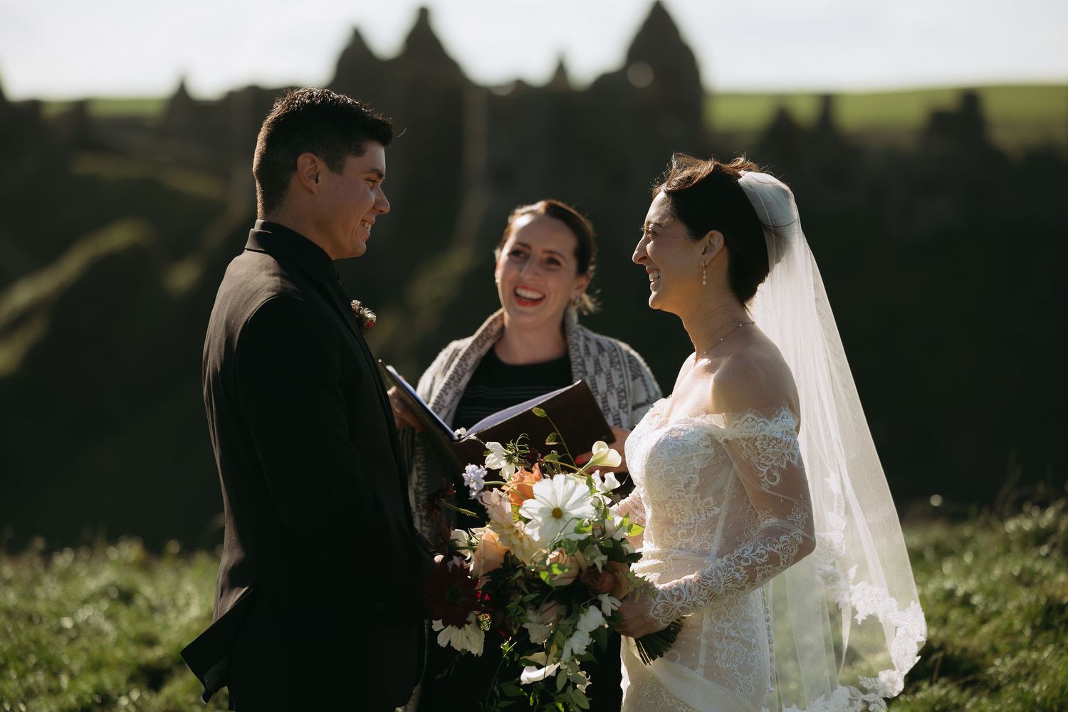 Bride and groom exchange vows outdoors with officiant holding bouquet, scenic green hills in background.
