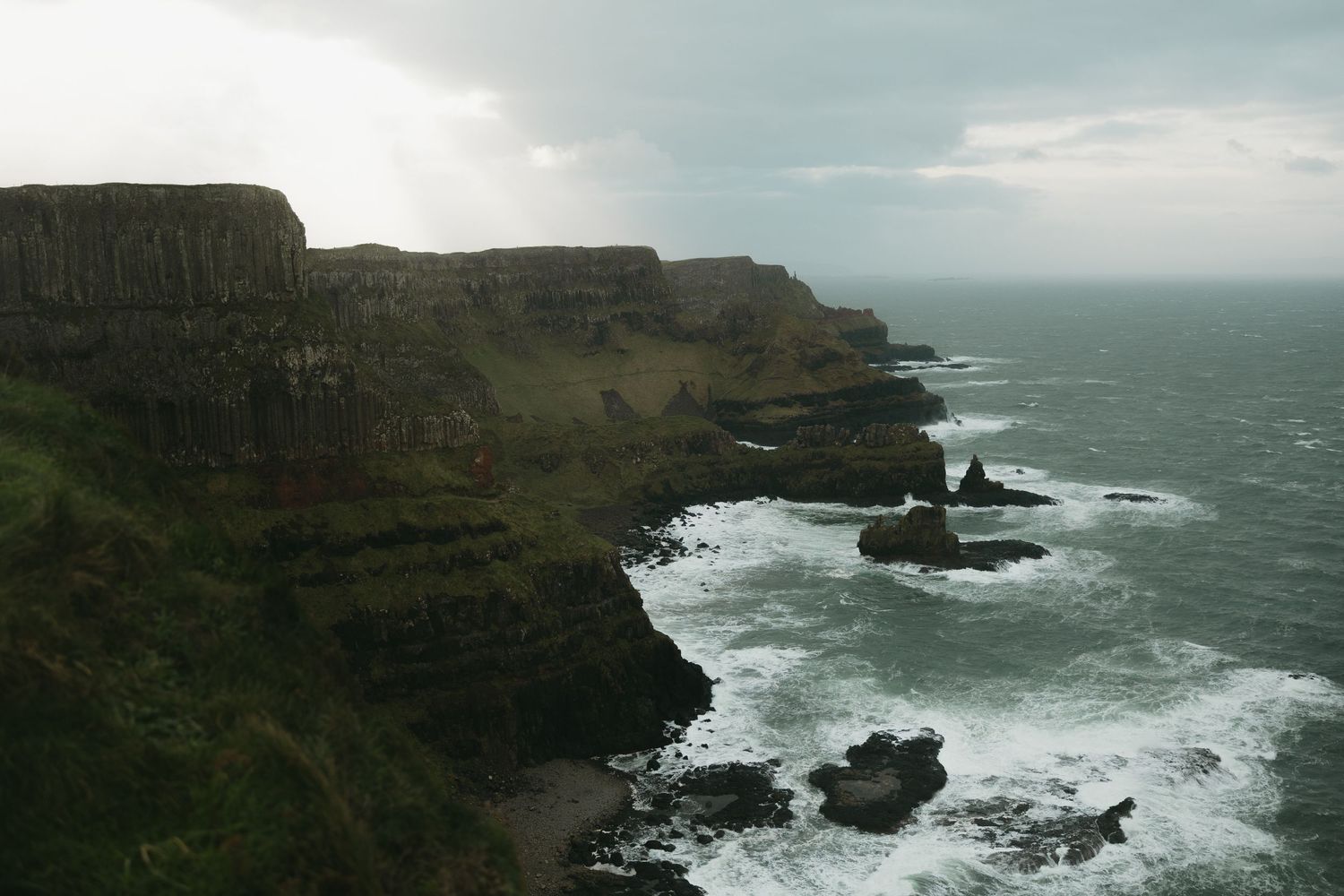 Dramatic rocky coastline with waves crashing against cliffs under a cloudy sky.