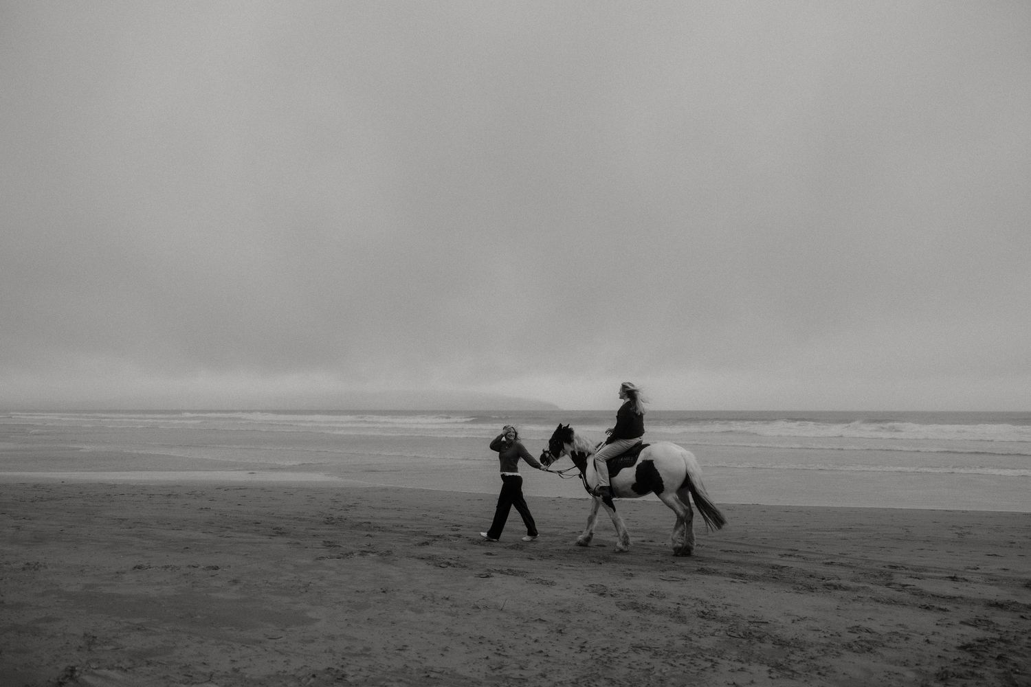 Black and white photo of a person leading a rider on a white horse along a misty beach shoreline.