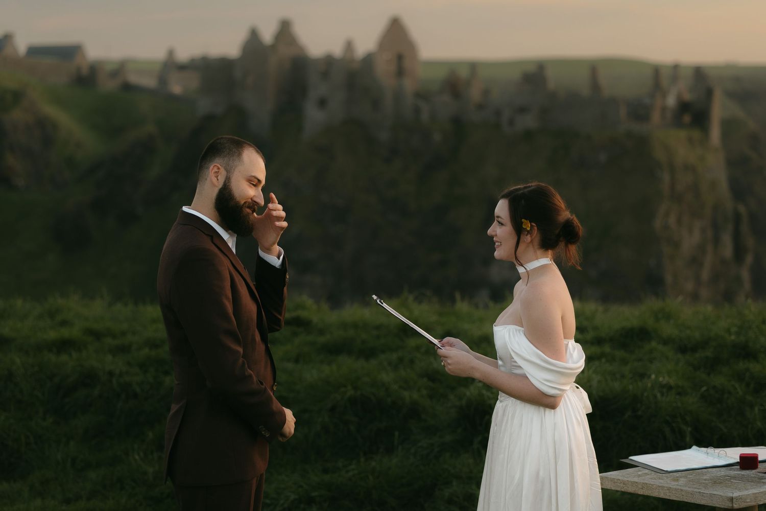 Couple exchanging vows outdoors at sunset with dramatic green cliffs and rock formations in the background.