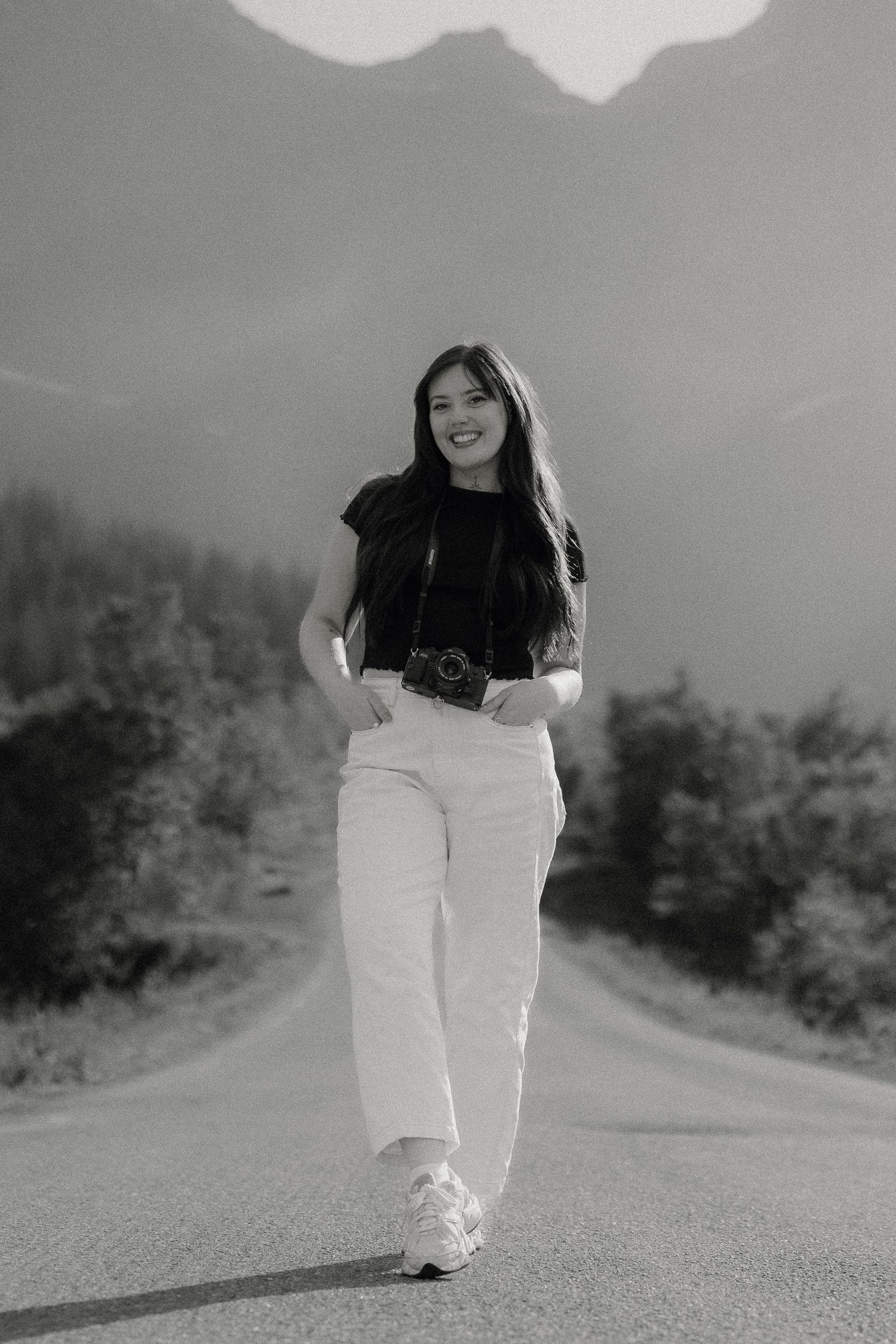 Black and white photo of a smiling woman walking on a road with mountains and trees in the background.