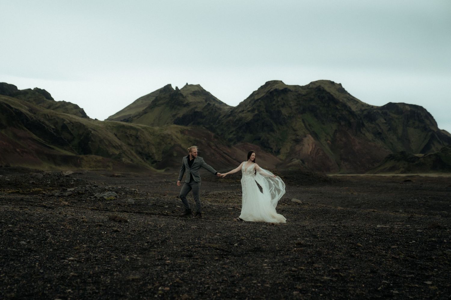A wedding couple dances together on black volcanic sand with mountains in the background.