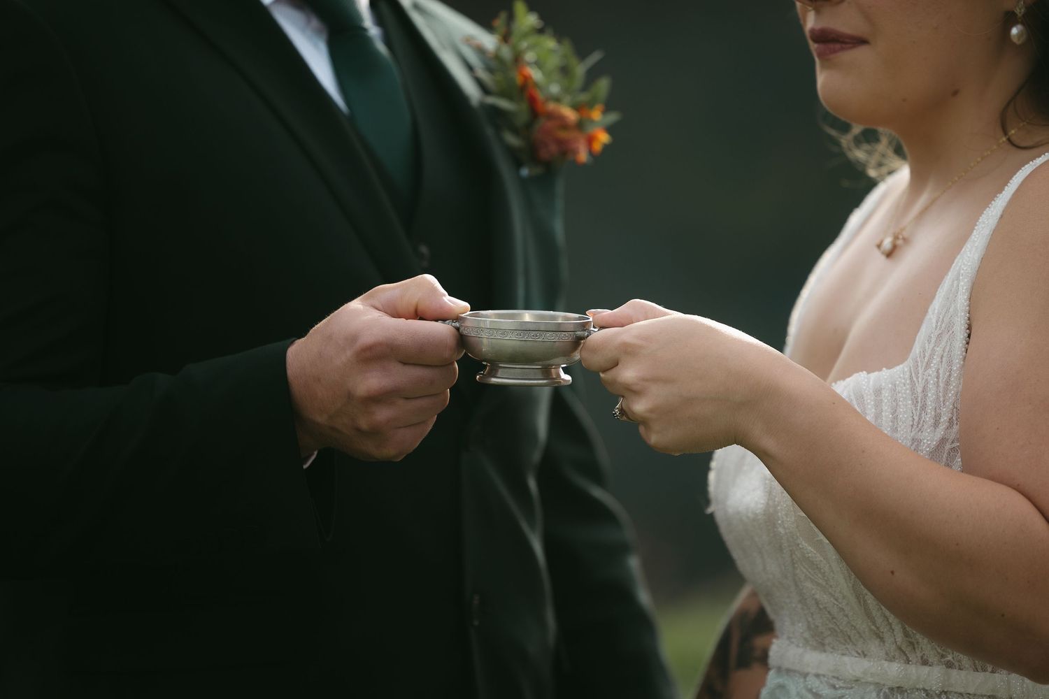 Bride and groom sharing a cup of tea during an outdoor wedding ceremony, dressed in white gown and dark suit.