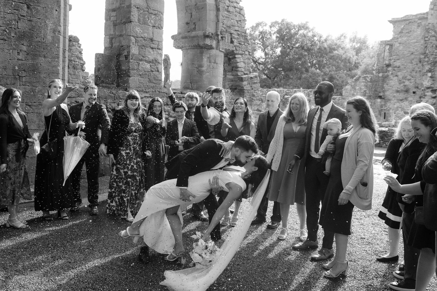 Black and white wedding photo of couple bowing to guests at an outdoor ceremony amid ancient stone columns.