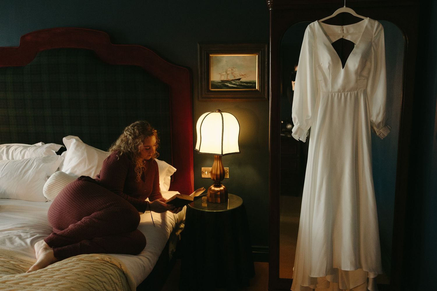 Bride sitting on bed beside lamp reviewing details, white wedding dress hanging nearby in dimly lit hotel room.